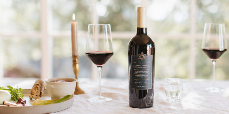 OneHope Wine's Kosher Cabernet bottle on table with 2 filled wine glasses and a plate of food