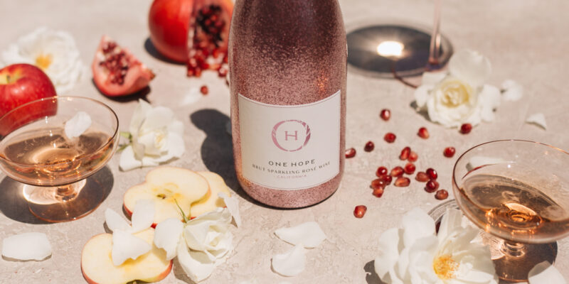 ONEHOPE Sparkling Rosé bottle with glasses of bubbly, pomegranate, apples, and white roses—perfect for a festive Galentine’s Night In celebration.